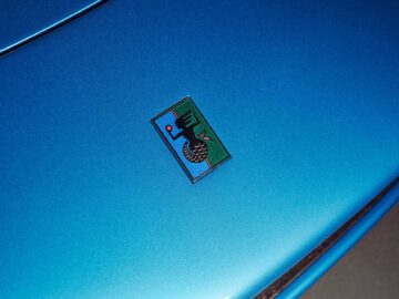 Close-up van de motorkap van een blauwe auto met een kleurrijk embleem met een haan en een band, symbool voor het Iso Rivolta-logo, dat doet denken aan het voortreffelijke vakmanschap dat te zien is in de Gordon Murray Automotive T.33 Spider.