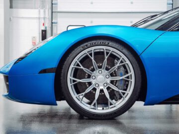 Close-up zijaanzicht van het voorwiel en spatbord van een blauwe sportwagen, met name de Gordon Murray Automotive T.33 Spider, met zilverkleurige velgen en Michelin-banden, in een garage.