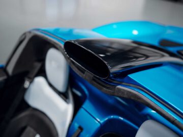 Close-up van de luchtinlaat van een blauwe Gordon Murray Automotive T.33 Spider, gelegen tussen de stoelen. Het interieur is voorzien van witte en zwarte bekleding, wat het aerodynamische ontwerp van de auto benadrukt.