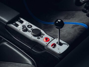 Close-up van de middenconsole van een auto met een handgeschakelde versnellingspook, verschillende knoppen en schakelaars, met een blauw veiligheidsharnas zichtbaar op de achtergrond, wat de ingewikkelde details van de Gordon Murray Automotive T.33 Spider benadrukt.