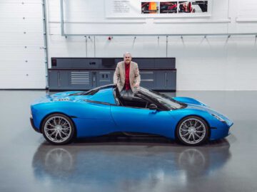 Een man in een beige blazer staat naast een blauwe Gordon Murray Automotive T.33 Spider in een ruime, goed verlichte garage.