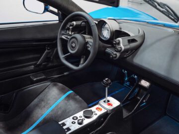 Interieur van een moderne sportwagen met een strak zwart en blauw ontwerp, een stuur met meerdere bedieningselementen, een gecentreerde versnellingspook en een digitaal instrumentenpaneel, wat de verfijning van de Gordon Murray Automotive T.33 Spider oproept.