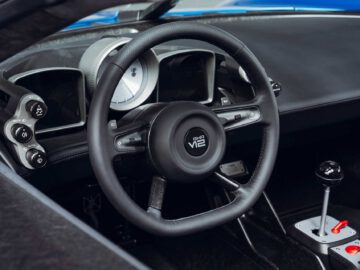 Close-up van het stuur en het dashboard van een Gordon Murray Automotive T.33 Spider, met een handmatige versnellingspook, verschillende draaiknoppen en bedieningsknoppen.