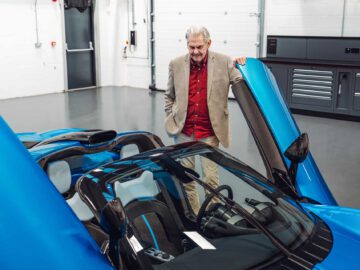 Een oudere man in een beige blazer en rood overhemd staat naast een open blauwe Gordon Murray Automotive T.33 Spider in een garage en kijkt er geïnteresseerd naar.