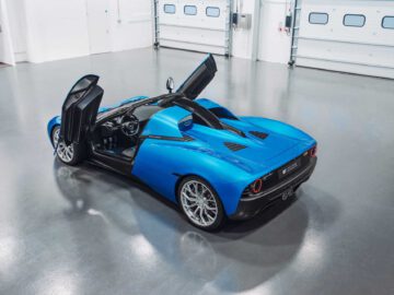 Een Gordon Murray Automotive T.33 Spider met open schaardeuren staat elegant in een ruime, strakke garage met witte muren en grote gesloten garagedeuren op de achtergrond.