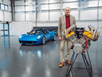 Een man in een blazer staat naast een zichtbare motor op een standaard, met een blauwe Gordon Murray Automotive T.33 Spider op de achtergrond in een garage.