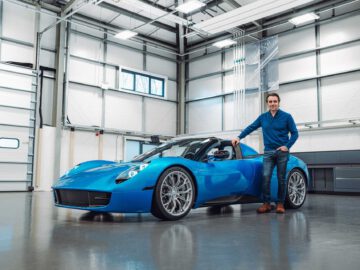 Een persoon staat naast een blauwe Gordon Murray Automotive T.33 Spider in een moderne garage met grote ramen en een hoog plafond.