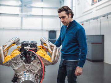 Een man in een blauwe trui en spijkerbroek onderzoekt een krachtige Gordon Murray Automotive T.33 Spider-motor in een industriële omgeving.