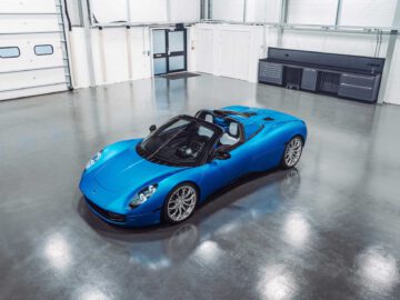 Een blauwe sportwagen, met name de gestroomlijnde Gordon Murray Automotive T.33 Spider, staat geparkeerd in een schone, ruime garage met witte muren, een gesloten garagedeur en zwarte opbergruimtes.