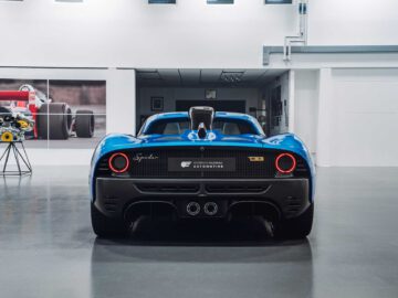 Achteraanzicht van een blauwe Gordon Murray Automotive T.33 Spider binnenshuis tentoongesteld voor een witte muur, met links een kunstwerk van een Formule 1-auto en een zonnebloem.