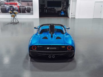 Een blauwe Gordon Murray Automotive T.33 Spider staat geparkeerd in een schone, moderne garage met een gepolijste vloer en witte muren. De achterkant van de auto is naar de camera gericht. Een ingelijste autofoto en een bloemstuk zijn zichtbaar.