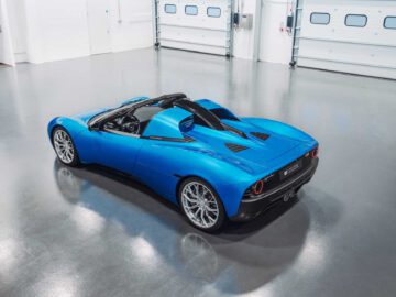 Een blauwe converteerbare Gordon Murray Automotive T.33 Spider-sportwagen staat binnen geparkeerd op een gepolijste betonnen vloer, met het dak naar beneden en garagedeuren op de achtergrond.