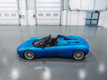 Een blauwe Gordon Murray Automotive T.33 Spider staat met de kap naar beneden geparkeerd in een moderne, strakke garage met witte muren en gesloten overheaddeuren.