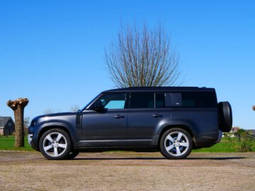 Een donkergekleurde Defender-SUV staat geparkeerd op een geplaveid terrein met op de achtergrond boomstronken en een open landschap onder een helderblauwe lucht.