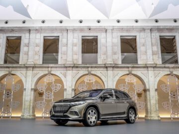 Een luxe Maybach SUV staat geparkeerd voor een gebouw met boogramen en een sierlijke gevel. Het plafond erboven heeft een geometrisch patroon.