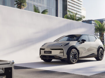 Buiten staat een moderne ZEEKR elektrische SUV, tegen een witte achtergrond met een strakke stedelijke omgeving op de achtergrond. Bomen en gebouwen zijn zichtbaar.