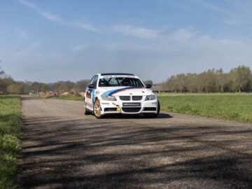 Een BMW 325i racewagen met sponsorlogo's rijdt over een verharde landweg, omgeven door groene velden en bomen onder een helderblauwe lucht.