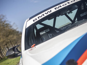 Close-up van de voorkant van een BMW 325i-raceauto, met de voorruit, het gedeeltelijke interieur en een 'BMW Motorsport'-banner bovenaan. De auto is voorzien van Sparco-racestoelen en een blauwe en rode streep aan de buitenkant.