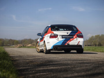 Een BMW 325i sedan met M Performance-logo staat geparkeerd op een landweg, voorzien van een racekleurstelling. Op een heldere dag staan bomen en gras langs de weg.