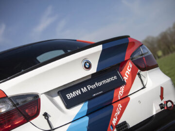 Close-up van de achterkant van een witte BMW M Performance-auto, die doet denken aan de iconische BMW 325i, met rode, blauwe en lichtblauwe racestrepen en branding op een heldere dag.