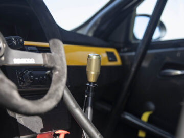 Close-up van het interieur van een BMW 325i met een stuur, dashboardbediening en een metalen versnellingspook op de voorgrond. Het interieur is voorzien van zwarte en gele accenten.