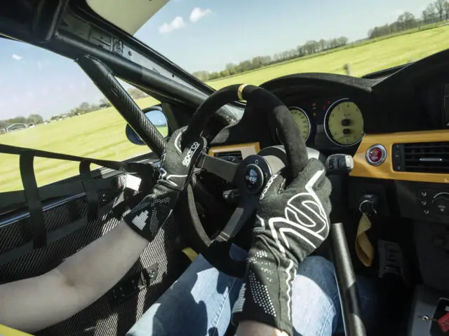 Binnenaanzicht van de cockpit van een BMW 325i raceauto met een persoon met racehandschoenen die het stuur bestuurt, instrumenten op het dashboard en een raceveiligheidsharnas zichtbaar, met een grasveld zichtbaar door de ramen.