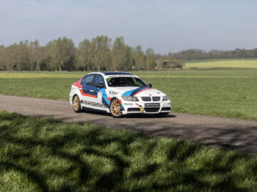 Een BMW 325i met het opschrift 'Performance' rijdt op een landelijke weg omringd door grasvelden en verre bomen onder een heldere hemel.