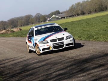 Een BMW 325i autosportwagen met afbeeldingen op de zijkant rijdt op een landelijke weg, omgeven door open velden en bomen op de achtergrond.