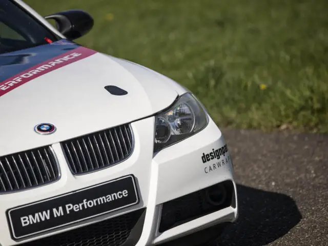 Close-up van de voorkant van een witte BMW M Performance-auto met designgraphics, geparkeerd op een asfaltoppervlak met een grasachtige achtergrond, die doet denken aan de iconische BMW 325i.