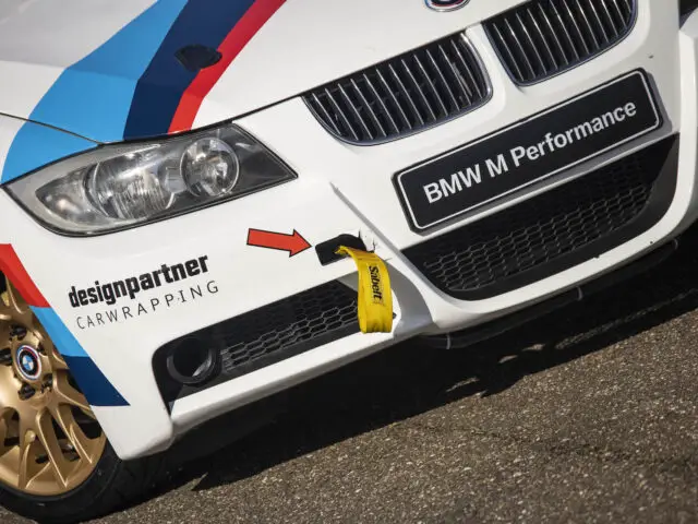 Close-up van een witte BMW 325i M Performance-auto met de voorbumper met sponsorstickers, waaronder een "designpartner Carwrapping"-logo en een gele sleepriem.