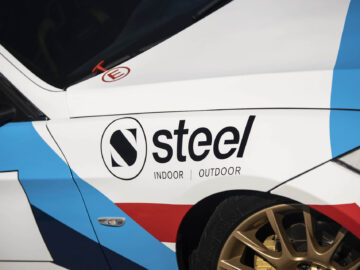 Close-up van een BMW 325i met het "Steel Indoor Outdoor"-logo op de zijkant, met een zwart "N"-symbool. De auto heeft een wit, blauw en rood kleurenschema met gouden wielen.