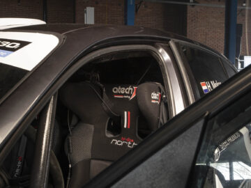 Uitzicht op het interieur van een raceauto, met een racestoel met het merk "Atech Racing" en harnasriemen. De deur aan de bestuurderszijde staat open en onthult de gedetailleerde zitopstelling van de BMW 130i Challenge.
