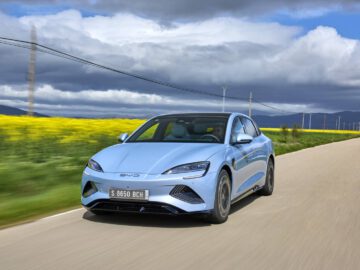 Een lichtblauwe BYD Seal sedan rijdt op een landelijke weg met aan de ene kant een veld met gele bloemen en heuvels in de verte onder een gedeeltelijk bewolkte hemel.