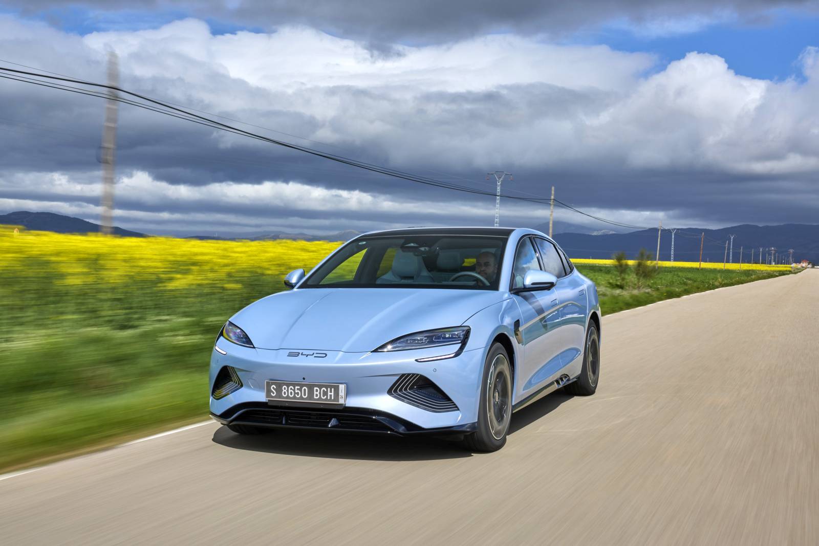 Een lichtblauwe BYD Seal sedan rijdt op een landelijke weg met aan de ene kant een veld met gele bloemen en heuvels in de verte onder een gedeeltelijk bewolkte hemel.