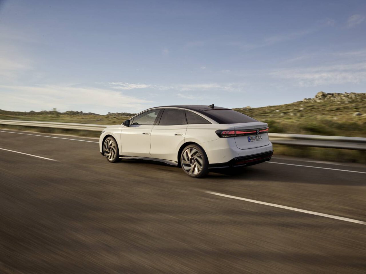 Een ID.7 met strak zwart dak rijdt op een verharde weg omzoomd door een metalen vangrail, met open grasvelden en een helderblauwe lucht op de achtergrond.