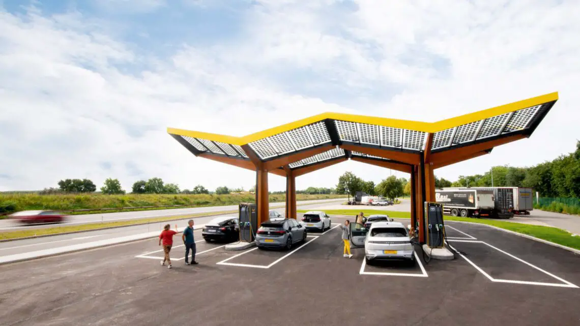 People charge electric vehicles on a clear day at a modern Fast Charging Station with rooftop solar panels near a highway.