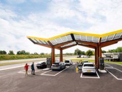 Mensen laden op een heldere dag elektrische voertuigen op bij een modern Snelladen-laadstation met dakbedekking met zonnepanelen nabij een snelweg.