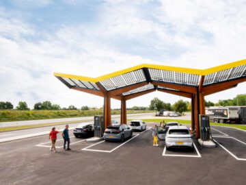 Mensen laden op een heldere dag elektrische voertuigen op bij een modern Snelladen-laadstation met dakbedekking met zonnepanelen nabij een snelweg.