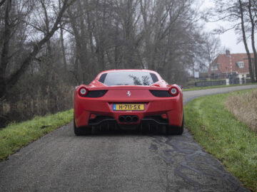 Op een smalle, bochtige weg omgeven door bomen en groen staat een rode Ferrari 458 Italia met Nederlands kenteken geparkeerd. Op de achtergrond zijn huizen zichtbaar.