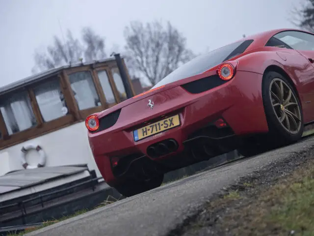 Een Ferrari 458 Italia-sportwagen staat geparkeerd bij een boot, van achteren gefotografeerd, met het kenteken zichtbaar.