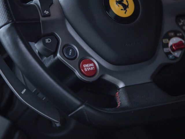 Close-up van het stuur van een auto met focus op de rode "Engine Start"-knop, een centraal embleem en bedieningselementen aan de linkerkant, typisch voor de luxueuze Ferrari 458 Italia.