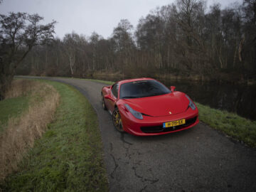 Een rode Ferrari 458 Italia-sportwagen staat geparkeerd op een smalle, bochtige weg naast een met gras bedekt gebied en een watermassa, omgeven door bladerloze bomen op een bewolkte dag.