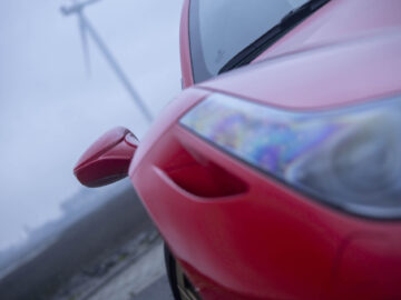 Close-up van het voorste gedeelte van een rode Ferrari 458 Italia, waarbij de nadruk ligt op de koplamp en de zijspiegel, met een windturbine zichtbaar op de mistige achtergrond.