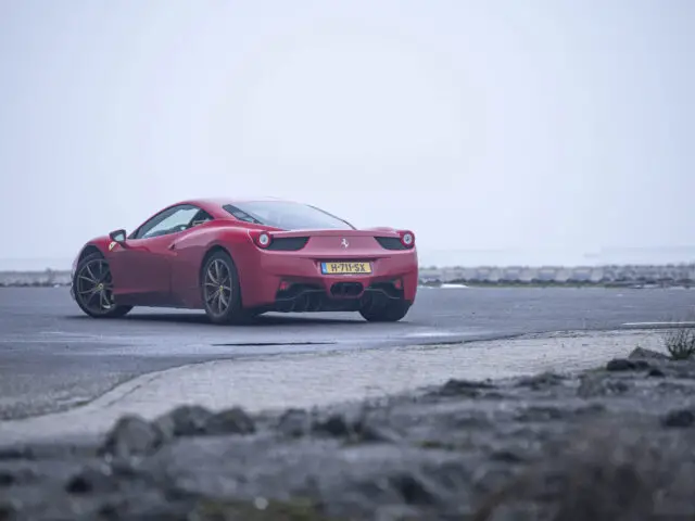 Een rode Ferrari 458 Italia staat geparkeerd op een mistige kustweg, weg van de camera. Het gebied is verlaten, met natte bestrating en verspreide rotsen.