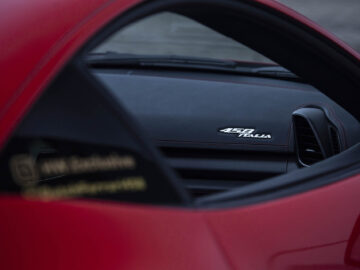 Close-up van het interieur van een Ferrari 458 Italia met het "458 Italia"-logo op het dashboard. De buitenkant van de auto is rood en op de ruit zijn stickers te zien met gebruikersnamen voor sociale media.