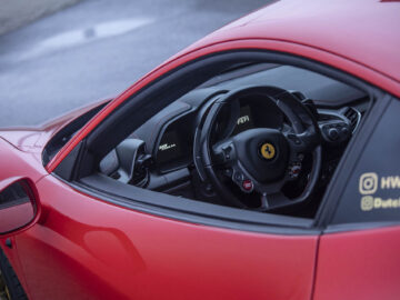 Binnenaanzicht van een rode Ferrari 458 Italia met een Ferrari-logo op het stuur, waarbij het dashboard en de bestuurdersstoel door een open raam zichtbaar zijn.