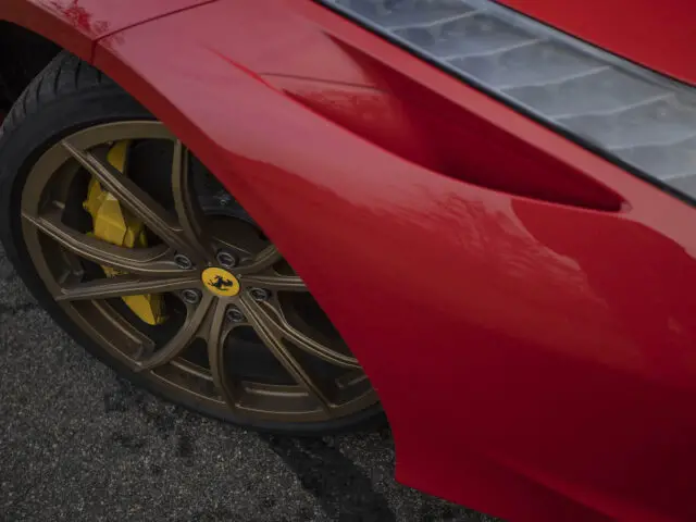 Close-up van het linkervoorwiel van een rode sportwagen van een Ferrari 458 Italia, met gouden velgen met een gele remklauw. De afbeelding toont ook een deel van de carrosserie van de auto.