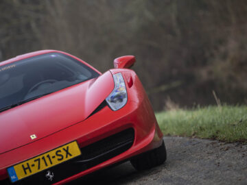 Close-up van een Ferrari 458 Italia met een zichtbaar kenteken "H-711-SX", geparkeerd op een weg nabij gras en bomen.