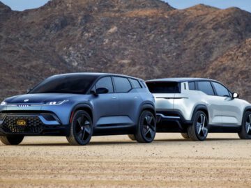 Twee elektrische SUV's, een donkergrijze en een witte Fisker Ocean, staan naast elkaar geparkeerd in een woestijnlandschap met bergen op de achtergrond.