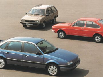 Op een vlakke, lege ondergrond staan drie klassieke auto's geparkeerd: een blauwe Volkswagen Passat sedan op de voorgrond, een rode tweedeurs coupé in het midden en een bruine stationwagen op de achtergrond.